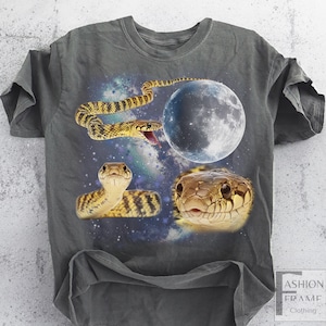May include: Dark gray t-shirt featuring a vibrant graphic of snakes, a full moon, and a starry galaxy background. The snakes are depicted in shades of yellow, brown, and tan. The shirt has a relaxed fit and short sleeves.