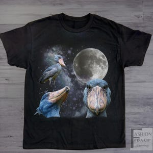 May include: Black t-shirt featuring a graphic design of a Shoebill stork with a full moon and starry background. The design includes three different views of the bird. The shirt has a crew neck and short sleeves.