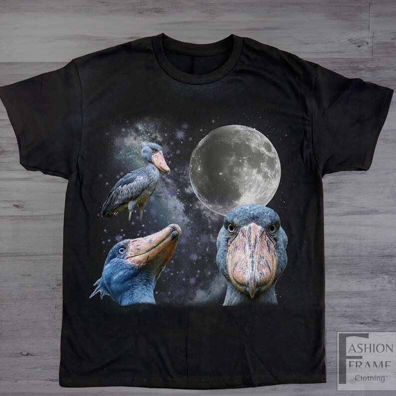 May include: Black t-shirt featuring a graphic design of a Shoebill stork with a full moon and starry background. The design includes three different views of the bird. The shirt has a crew neck and short sleeves.