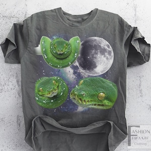 May include: Dark gray t-shirt featuring a graphic design of three green snakes and a full moon against a starry background. The snakes are detailed with white spots and yellow eyes. The shirt has a crew neck and short sleeves.