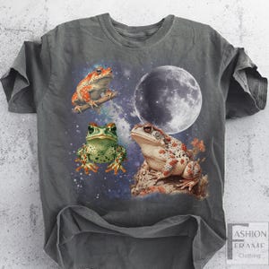 May include: Dark gray t-shirt featuring a whimsical design of three frogs against a starry night sky with a full moon. The frogs are in shades of green, red, and brown. The shirt has a relaxed fit and the words "FASHION FRAME CLOTHING" are printed on the bottom right.