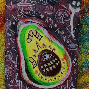 Könnte beinhalten: Ein farbenfrohes Gemälde einer Avocado mit einem Comic-Gesicht. Die Avocado ist grün mit einer gelben Mitte und einem braunen Gesicht mit einem einzigen Auge und einem breiten, zahnigen Grinsen. Der Hintergrund ist dunkel mit weißen und roten Kritzeleien.