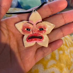 May include: A handmade, flower-shaped pendant with a face painted on it. The pendant is cream-colored with pink and red accents, and has a black cord. The face has red lips and dark eyes. The pendant is held in a hand.
