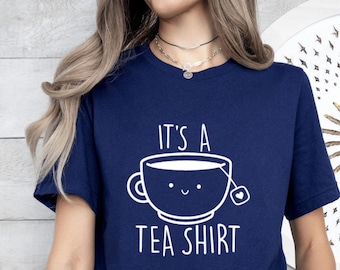 May include: Navy blue t-shirt with the text "IT'S A TEA SHIRT" and a smiling teacup graphic in white. The graphic includes a teabag. The shirt is short-sleeved.