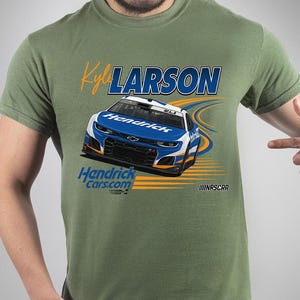 May include: Olive green t-shirt featuring a graphic of a race car with the name "Kyle Larson" and the text "HendrickCars.com". The car is blue and white with orange accents. The NASCAR logo is visible.