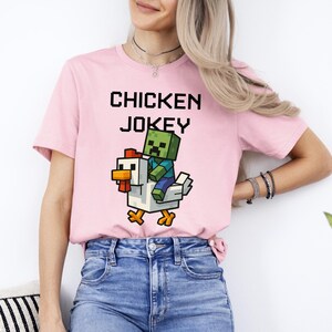 May include: A light pink t-shirt with the words "CHICKEN JOKEY" in pixelated black letters. Below the text is a graphic of a pixelated zombie riding a pixelated chicken. The chicken is white with orange and red accents.