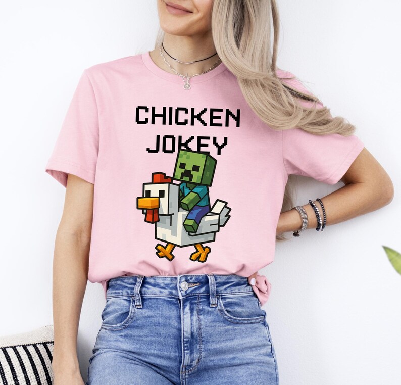 May include: A light pink t-shirt with the words "CHICKEN JOKEY" in pixelated black letters. Below the text is a graphic of a pixelated zombie riding a pixelated chicken. The chicken is white with orange and red accents.