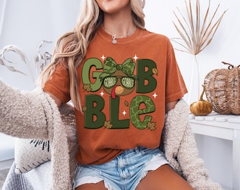 May include: An orange t-shirt with the word "GOBBLE" in green letters. The "O"s are turkey heads wearing sunglasses and a bow tie. The shirt is paired with denim shorts, perfect for Thanksgiving.
