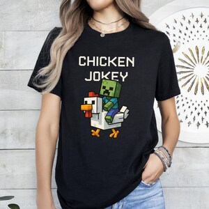 May include: Black t-shirt with the text "CHICKEN JOKEY" in white block letters. Below the text is a pixelated graphic of a zombie riding a chicken. The chicken is white with orange and yellow accents.