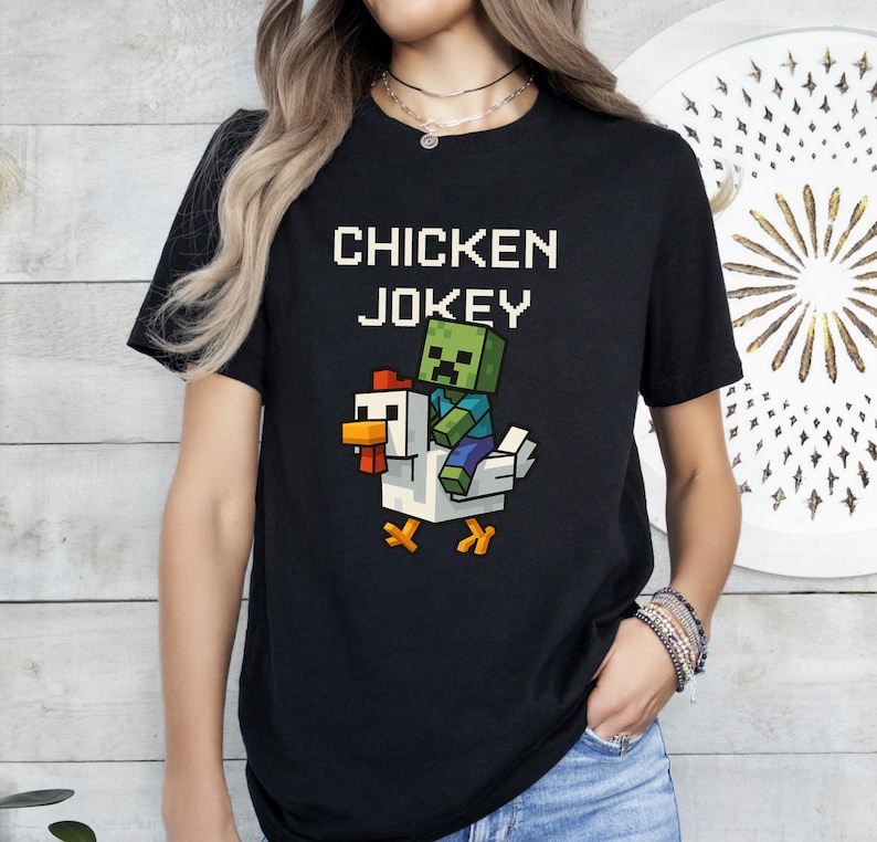 May include: Black t-shirt with the text "CHICKEN JOKEY" in white block letters. Below the text is a pixelated graphic of a zombie riding a chicken. The chicken is white with orange and yellow accents.