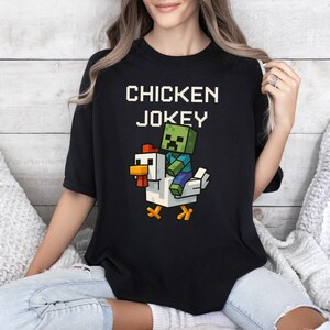 May include: Black t-shirt with the text "CHICKEN JOKEY" in white block letters. A pixelated green zombie character rides a white chicken with orange beak and feet. The design is centered on the shirt.