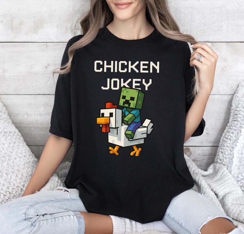 May include: Black t-shirt with the text "CHICKEN JOKEY" in white block letters. A pixelated green zombie character rides a white chicken with orange beak and feet. The design is centered on the shirt.