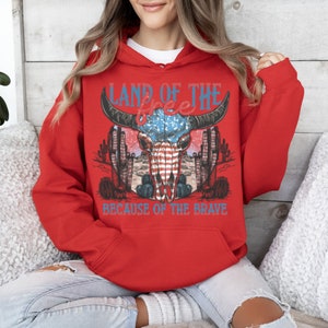 May include: Red hooded sweatshirt with a graphic print of a bull skull with an American flag design. The text "Land of the Free because of the Brave" is printed below the skull.