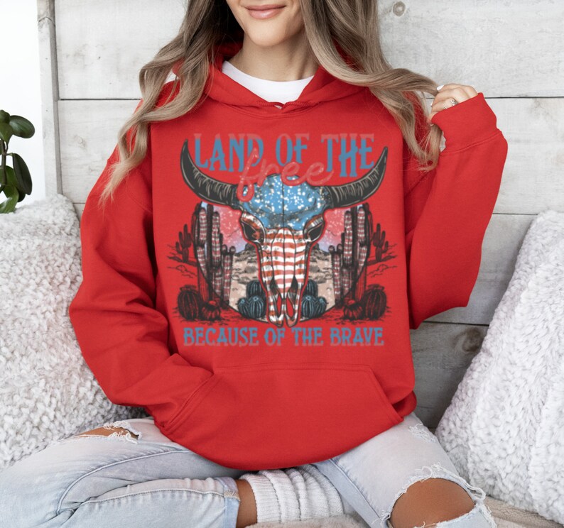 May include: Red hooded sweatshirt with a graphic print of a bull skull with an American flag design. The text "Land of the Free because of the Brave" is printed below the skull.