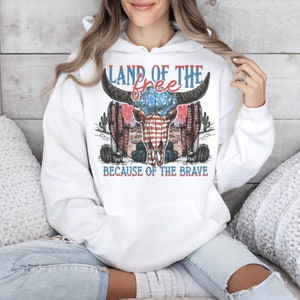 May include: White hooded sweatshirt with a vintage-style graphic of a skull with an American flag design. The text "Land of the free because of the brave" is printed on the sweatshirt.