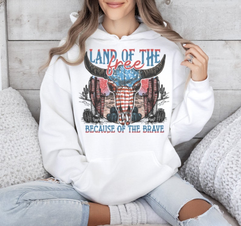 May include: White hooded sweatshirt with a vintage-style graphic of a skull with an American flag design. The text "Land of the free because of the brave" is printed on the sweatshirt.