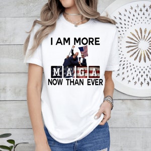 May include: White t-shirt with the text "I am more MAGA now than ever" printed on it. The text is in black and the letters MAGA are in red, white, and blue boxes. There is a picture of four people holding an American flag above the text.