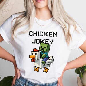 May include: White t-shirt with the text "CHICKEN JOKEY" in black pixelated font. The graphic features a pixelated zombie riding a pixelated chicken. The zombie is green and blue, and the chicken is white, yellow, and red.