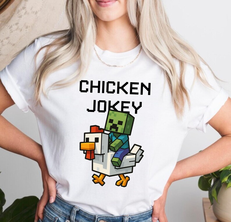 May include: White t-shirt with the text "CHICKEN JOKEY" in black pixelated font. The graphic features a pixelated zombie riding a pixelated chicken. The zombie is green and blue, and the chicken is white, yellow, and red.