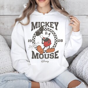 May include: White hooded sweatshirt featuring a distressed graphic of Mickey Mouse. The design includes the words "Mickey Mouse" and the year "1928" in a vintage style. The Disney logo is at the bottom of the graphic.