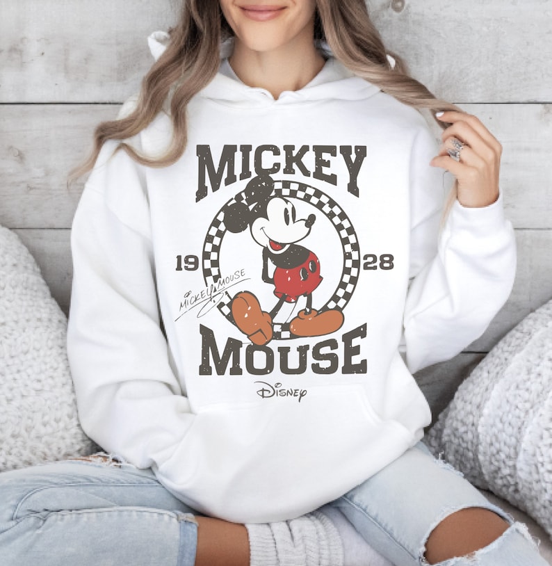 May include: White hooded sweatshirt featuring a distressed graphic of Mickey Mouse. The design includes the words "Mickey Mouse" and the year "1928" in a vintage style. The Disney logo is at the bottom of the graphic.