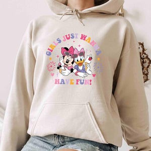 May include: A beige hoodie with a graphic print of Minnie Mouse and Daisy Duck wearing sunglasses and holding ice cream cones. The text "Girls Just Wanna Have Fun!" is printed in a rainbow arc above the characters.
