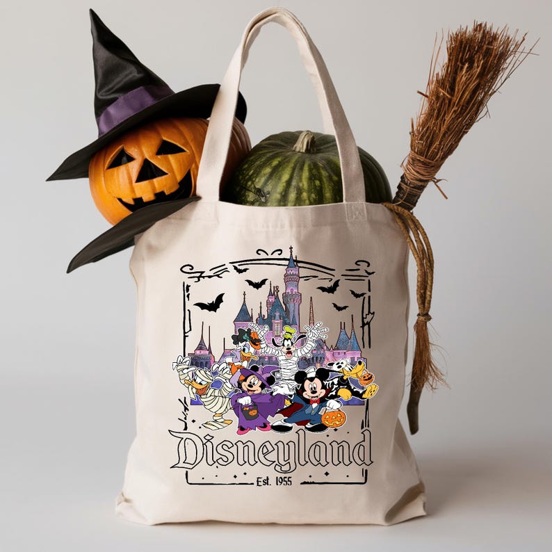 May include: A cream-colored tote bag featuring a Disneyland Halloween design with Mickey Mouse and friends in costume. A carved pumpkin, witch hat, and green pumpkin are inside the bag, with a broomstick on the side. The text "Disneyland Est. 1955" is printed on the bag.