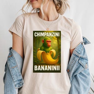 May include: Beige t-shirt featuring a graphic of a green monkey with a red face emerging from a banana peel. The text "CHIMPANZINI" is above the image, and "BANANINII" is below.