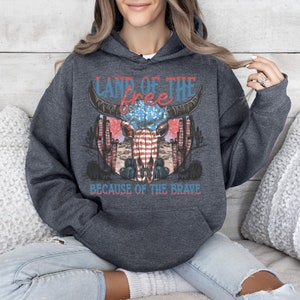 May include: A dark gray hooded sweatshirt with a graphic print of a skull with an American flag design. The text "Land of the free because of the brave" is printed below the skull.