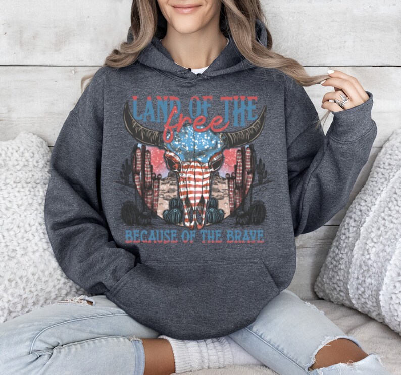 May include: A dark gray hooded sweatshirt with a graphic print of a skull with an American flag design. The text "Land of the free because of the brave" is printed below the skull.