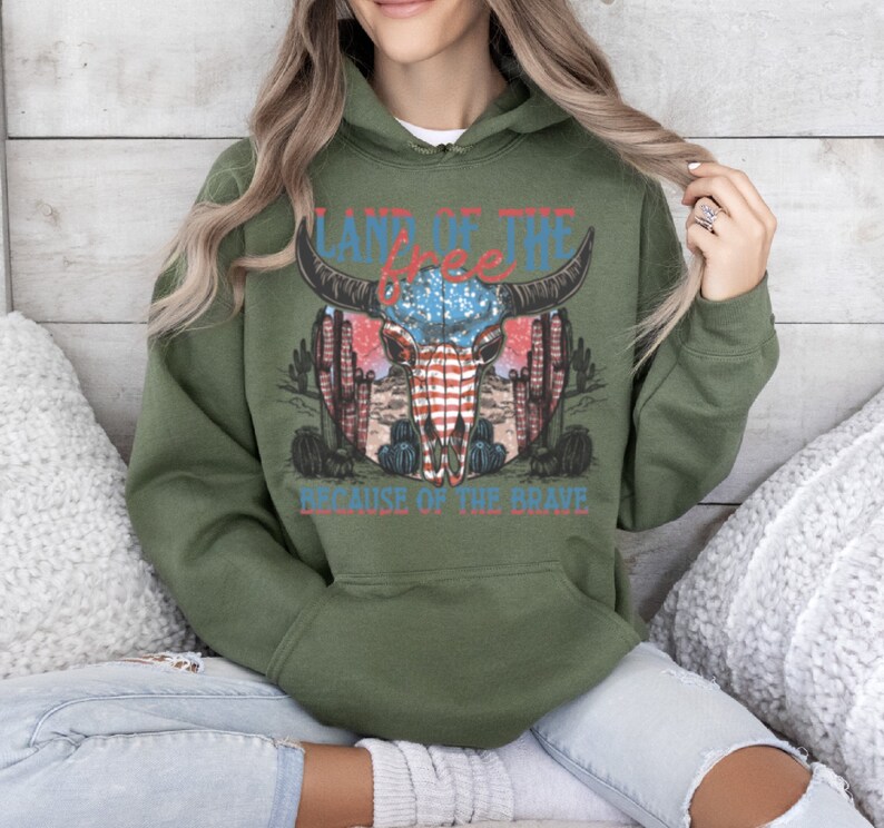 May include: Green hooded sweatshirt with a graphic print of a skull with an American flag design. The text "Land of the Free Because of the Brave" is printed below the skull.