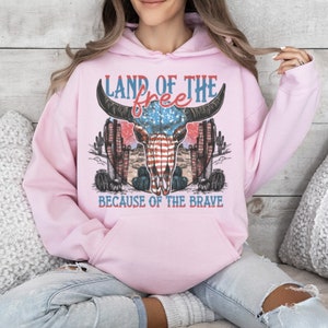 May include: Pink hoodie with a graphic of a bull skull with an American flag design. The text "Land of the free because of the brave" is printed below the skull.