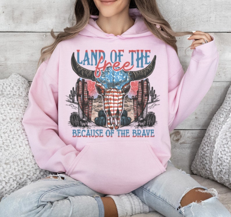 May include: Pink hoodie with a graphic of a bull skull with an American flag design. The text "Land of the free because of the brave" is printed below the skull.
