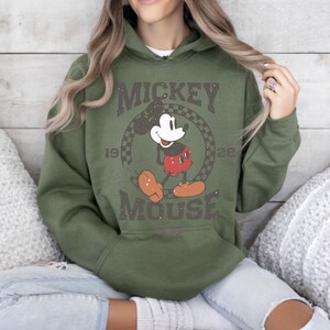 May include: Olive green hoodie featuring a distressed graphic of Mickey Mouse. The design includes the words "Mickey Mouse" and the years "1928" and a checkered circle. The hoodie has a drawstring hood and a kangaroo pocket.