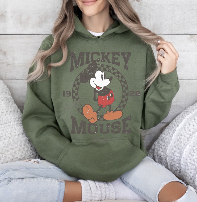 May include: Olive green hoodie featuring a distressed graphic of Mickey Mouse. The design includes the words "Mickey Mouse" and the years "1928" and a checkered circle. The hoodie has a drawstring hood and a kangaroo pocket.