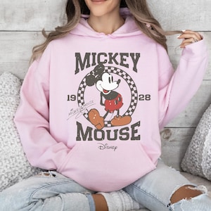 May include: Light pink hoodie featuring a distressed graphic of Mickey Mouse. The design includes the words "Mickey Mouse" and the years "1928" and "1928". The Disney logo is at the bottom. The hoodie has a drawstring hood and a front pocket.