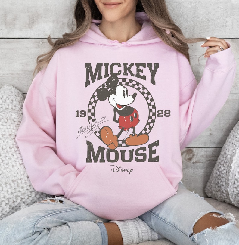 May include: Light pink hoodie featuring a distressed graphic of Mickey Mouse. The design includes the words "Mickey Mouse" and the years "1928" and "1928". The Disney logo is at the bottom. The hoodie has a drawstring hood and a front pocket.