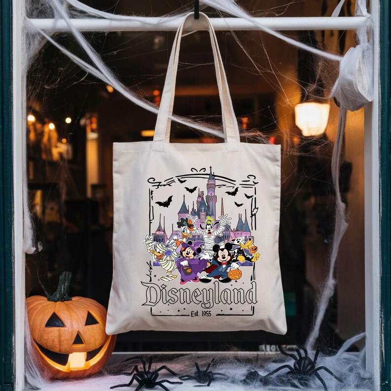 May include: Off-white tote bag featuring a Halloween-themed Disneyland graphic. The design includes Disney characters in costumes, a castle, bats, and the text "Disneyland Est. 1955". A carved pumpkin and spider decorations are in the background.