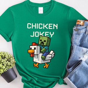 May include: A green t-shirt with the text "CHICKEN JOKEY" in white pixelated letters. The graphic shows a pixelated zombie riding a white chicken with orange accents. The shirt is paired with blue jeans.