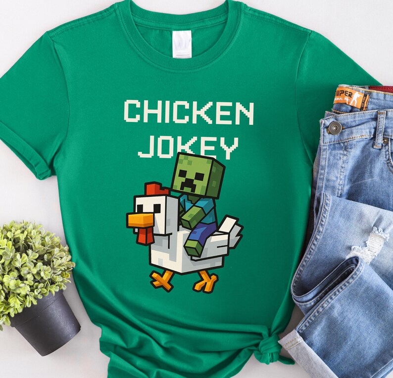 May include: A green t-shirt with the text "CHICKEN JOKEY" in white pixelated letters. The graphic shows a pixelated zombie riding a white chicken with orange accents. The shirt is paired with blue jeans.