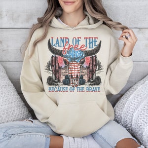 May include: A beige hoodie with a graphic print of a bull skull with an American flag design. The text "Land of the free because of the brave" is printed below the skull.