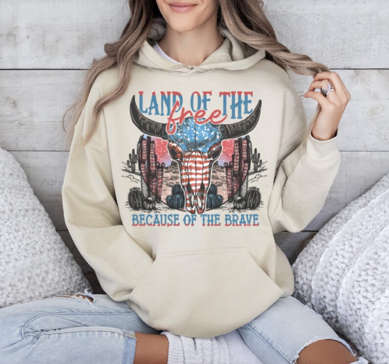 May include: A beige hoodie with a graphic print of a bull skull with an American flag design. The text "Land of the free because of the brave" is printed below the skull.