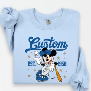 May include: Light blue sweatshirt featuring a baseball-themed Mickey Mouse graphic. The design includes the word "Custom" in a script font, the year "1958", and stars. Mickey is wearing a baseball uniform and holding a bat.