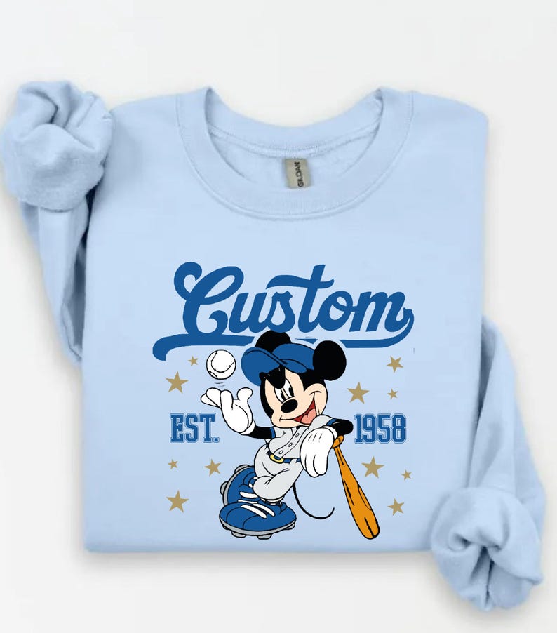 May include: Light blue sweatshirt featuring a baseball-themed Mickey Mouse graphic. The design includes the word "Custom" in a script font, the year "1958", and stars. Mickey is wearing a baseball uniform and holding a bat.