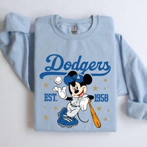May include: Pale blue sweatshirt with a blue "Dodgers" script and a Mickey Mouse graphic in a baseball uniform. Mickey is holding a bat and ball. The text "EST. 1958" is also visible on the sweatshirt.
