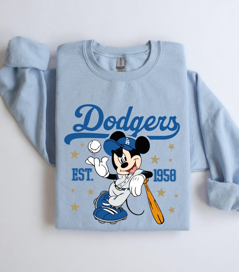 May include: Pale blue sweatshirt with a blue "Dodgers" script and a Mickey Mouse graphic in a baseball uniform. Mickey is holding a bat and ball. The text "EST. 1958" is also visible on the sweatshirt.