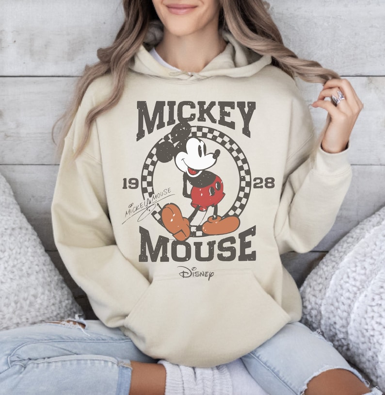 May include: Beige hoodie featuring a distressed graphic of Mickey Mouse. The design includes the text "Mickey Mouse" and the year "1928" within a checkered circle. The Disney logo is at the bottom of the graphic.