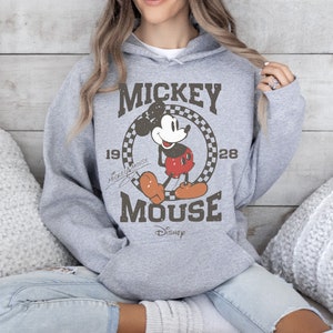 May include: A gray hooded sweatshirt featuring a vintage Mickey Mouse graphic. The design includes the words "Mickey Mouse" and the year "1928" in a circular frame. The Disney logo is at the bottom of the graphic.