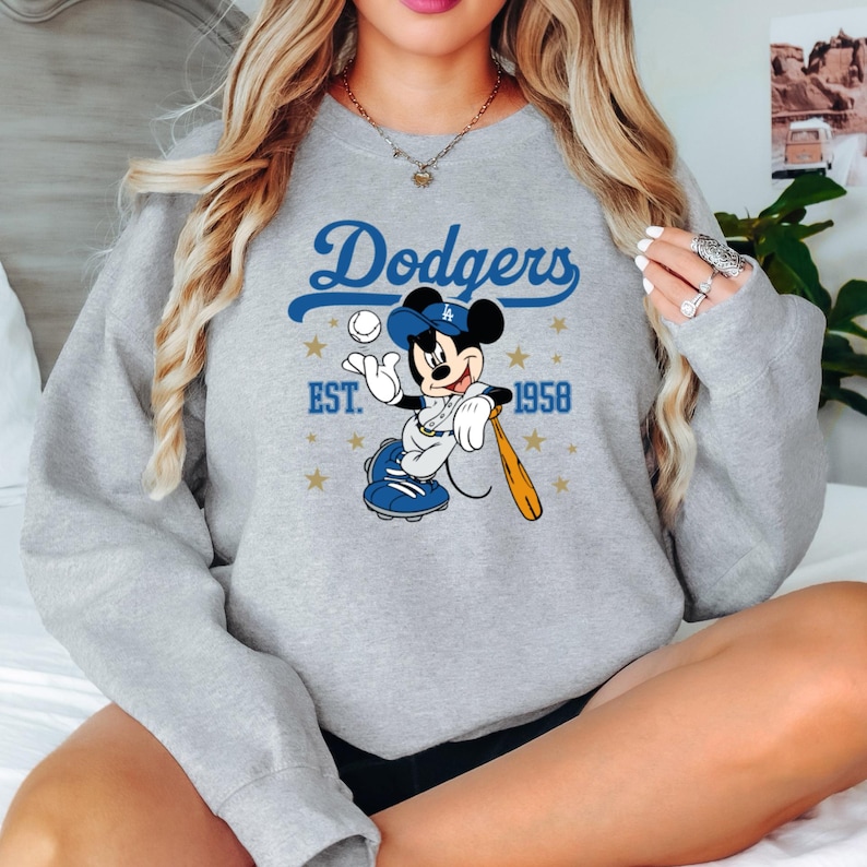 May include: A light grey sweatshirt with a Mickey Mouse baseball graphic. The design includes the word "Dodgers" in blue script, Mickey in a baseball uniform, a baseball, and a bat. The text "EST. 1958" is also included, with gold stars.