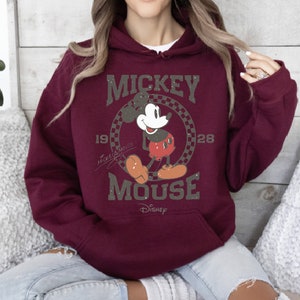 May include: Burgundy hoodie featuring a graphic of Mickey Mouse. The design includes the words "Mickey Mouse" and the years "1928" and "19". The Disney logo is at the bottom. The hoodie has a front pocket and a drawstring hood.